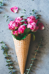 Still life of a bouquet of pink roses and eucalyptus inside a waffle cone on a grey textured background. The concept of gift and celebration. Flat lay, top view.
