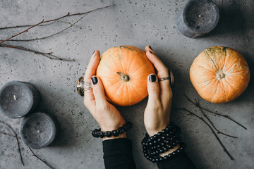 Witch's hands holding small pumpkin over grey background surrounded black candles and dried...