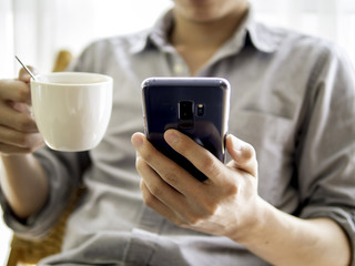 close up of Asian man relaxing with holding a cup of coffee and using a smartphone for his working, planning, scheduling or playing games, entertainment, social media networking or online payment,