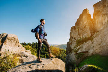 A traveler man standing on the rocks and holding backpack and binoculars over the blue sky background