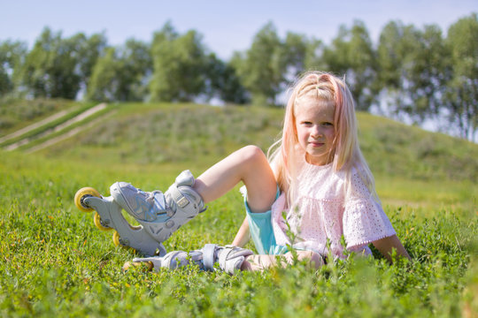 Cute little blonde girl sitting on green grass in roller skates and looking at camera - leisure, childhood, outdoor games and sport concept
