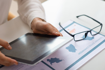 Cropped view of expert analyzing sales graphs. Close up of female using tablet at desk with paper charts and glasses. Business report concept