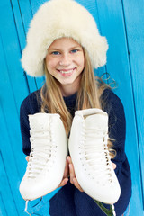 Cheerful little girl in warm sweater and hat keeps figure skates. Blue wooden background.