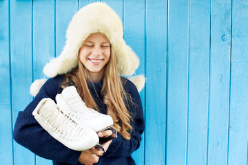 Cheerful little girl in warm sweater and hat keeps figure skates. Blue wooden background.