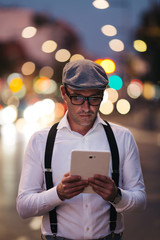 Fashionable retro dressed man with cap, suspenders and eyeglasses standing on city street and using tablet. City lights on sunset.