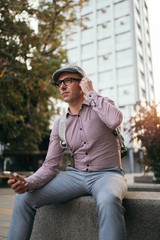 Fashionable retro dressed man with cap and eyeglasses sitting on city street and listening music with earphones