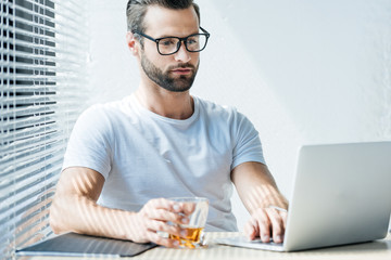 Fototapeta na wymiar brunet man holding glass of whiskey and working with laptop