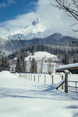 Winterlandschaft mit verschneiter Wiese, Holzzaun, Bergen und blauem Himmel