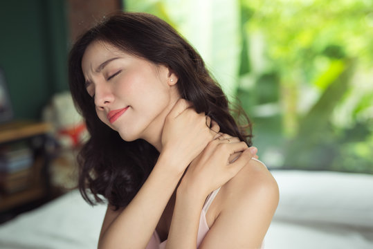 Young asian woman suffering from neck pain while sitting on bed at home