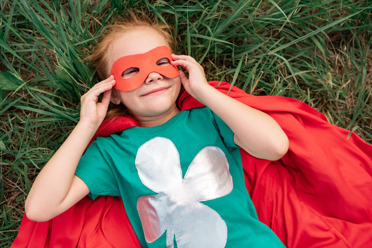 overhead view of kid in red superhero cape and mask lying on green grass