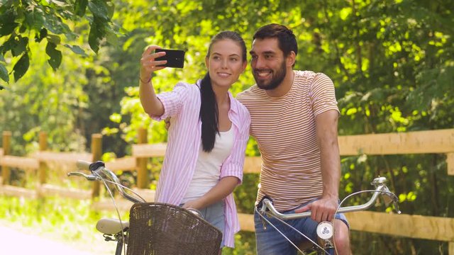 people, technology and lifestyle concept - happy couple with bicycles taking selfie by smartphone at summer park