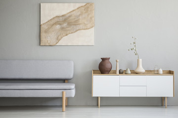 Real photo of a simple living room interior with a natural painting on the wall and gray sofa next...