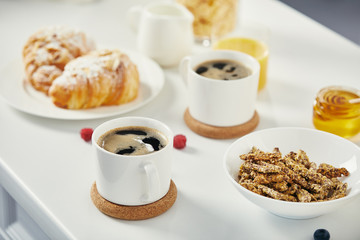 close up view of cups of coffee, croissants and raspberries for breakfast on white surface