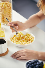 partial view of woman with spoon sitting at table with corn flakes and cup of coffee for breakfast