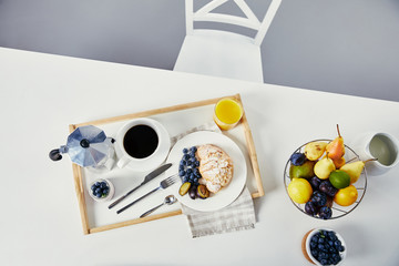 top view of croissant with fresh blueberries and plum pieces, glass of juice, cup of coffee and yogurt for breakfast on wooden tray on white tabletop