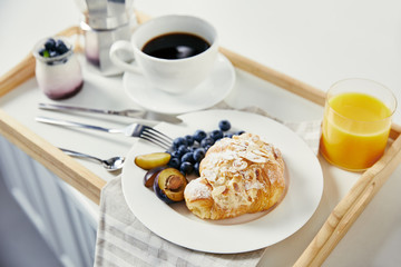close up view of croissant with fresh blueberries and plum pieces, glass of juice, cup of coffee and yogurt for breakfast on wooden tray on white tabletop