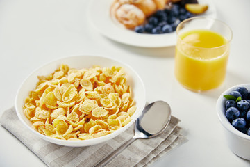 close up view of corn flakes in bowl, glass of juice and croissant with blueberries and plum pieces for breakfast on white tabletop
