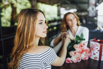 Mom's birthday! Daughter presents a gift and a bunch of roses on a summer terrace cafe in casual clothes. They are happy.