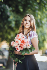 Portrait of a beautiful young blond girl standing with a bunch of roses on a blurry background in casual clothing. She is happy and smiling.