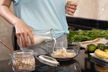 Crop woman pouring fresh milk into bowl with oatmeal and sweet banana while preparing breakfast in kitchen