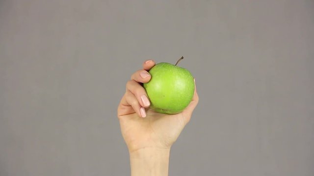 female hand holds an green apple on a gray background, rises from the bottom of the frame