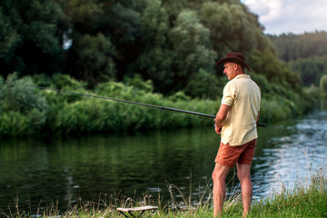 A fisherman in shorts, a hat and a T-shirt is fishing on the shore of the lake. Fishing, hobbies, recreation