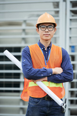 Young Asian man in waistcoat and hardhat holding blueprint and keeping arms crossed while standing...