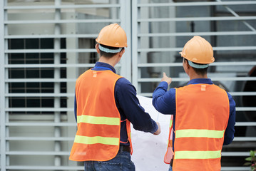 Back view of men in hardhats and waistcoats holding draft and pointing at building while standing...