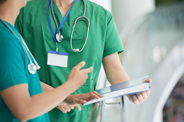 Two men in scrubs holding folder with medical records and discussing them while standing on blurred background of medical hall