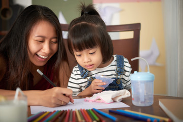 Adult Asian woman sitting at table with little daughter and drawing together on paper with colored pencils having fun 