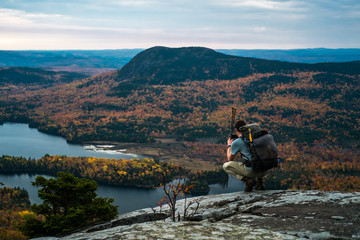 A man kneels on a mountain in Maine overlooking a lake and fall foliage while hiking the...