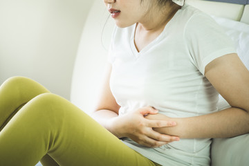 Asian young woman suffering from abdominal pain while sitting on bed at home