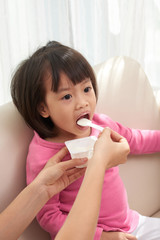 Crop hands of woman giving spoon with dairy yogurt to little Asian girl sitting on sofa in sunlight