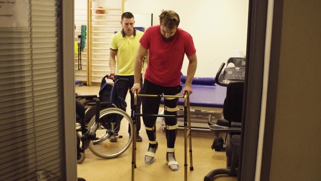 Young disabled man in orthosis walking holding a walking frame in the rehabilitation clinic. Doctor walking after him with wheelchair