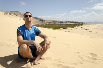 A portrait of a middle aged man in sunglasses sitting cross leged on the beach