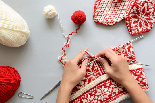 Girl knits red and white Norwegian jacquard hat knitting needles on gray wooden background. Process of knitting. Top view. Flat lay