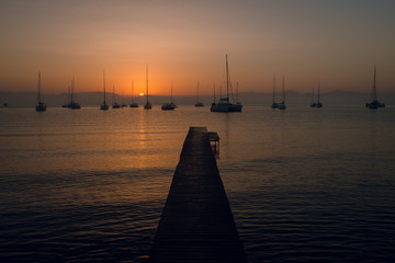 Fototapeta premium Scenic view of wooden pier and yachts at the calm sea on beautiful pink and orange sunset. Mountains on background. Dusk