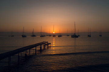 Obraz premium Wooden pier, yachts and boats at the calm sea bay with mountains on background at beautiful orange sunset. Dusk. Greece