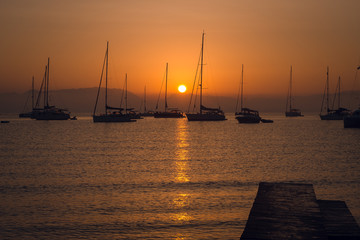 Sailing boats at the calm sea bay with mountains on background at beautiful scenic orange sunset. Dusk. Greece