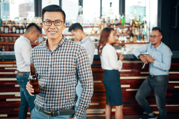 Adult Asian man standing with bottle of beer in bar and looking at camera with friends and colleagues on background 