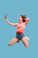 Fototapeta na wymiar always on mobile. Full length of pretty young woman taking phone while jumping against blue studio background. Mobile, motion, movement, business concepts