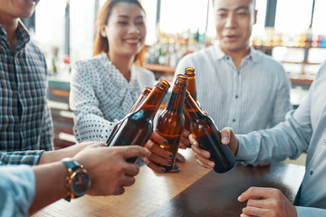 Crop people gathering together and clinking with beer bottles toasting for success in bar