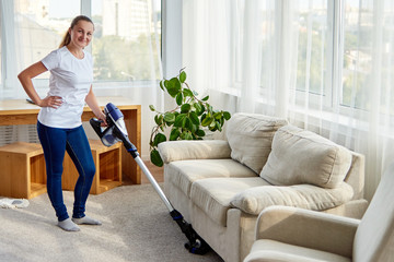 Full body portrait of young woman in white shirt and jeans cleaning carpet with vacuum cleaner in...