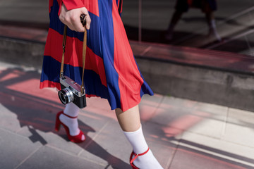 Obraz premium Elegant lady wearing stylish red skirt and shoes holds in hands vintage photo camera. Low angle view of her shapely legs. Selective focus