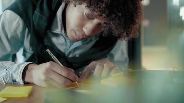 Close up of african young male student or creative start up businessman concentratedly writes notes on sticky multicolored stickers using pen leaning over desk brainstorming ideas and concepts slow mo