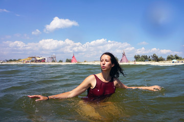 Young brunette girl in red swimsuit staring into the distance during swimming in a sea, with travel camp and teepee on a beach background and blue cloudy sky. Copyspace.