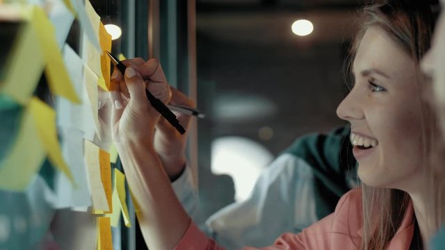 Cheerful european woman writing notes on sticky multicolored stickers at transparent glass wall multiethnic creative business team brainstorming analyzes concept planning strategy of start up project