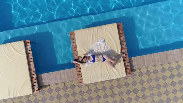 enjoying summer, aerial view of young girl in swimsuit lies on the terrace near pool with blue beautiful water