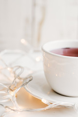 Vertical photo of red hibiscus tea in a white cup and small yellow christmas lights for home decoration on wooden table. Tea break