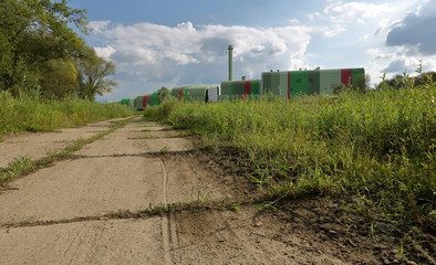 Polna droga, trawiasta łąka z prawej, za nią kolorowy budynek spalarni śmieci w Krakowie, Polska, małe chmurki na niebie - obrazy, fototapety, plakaty